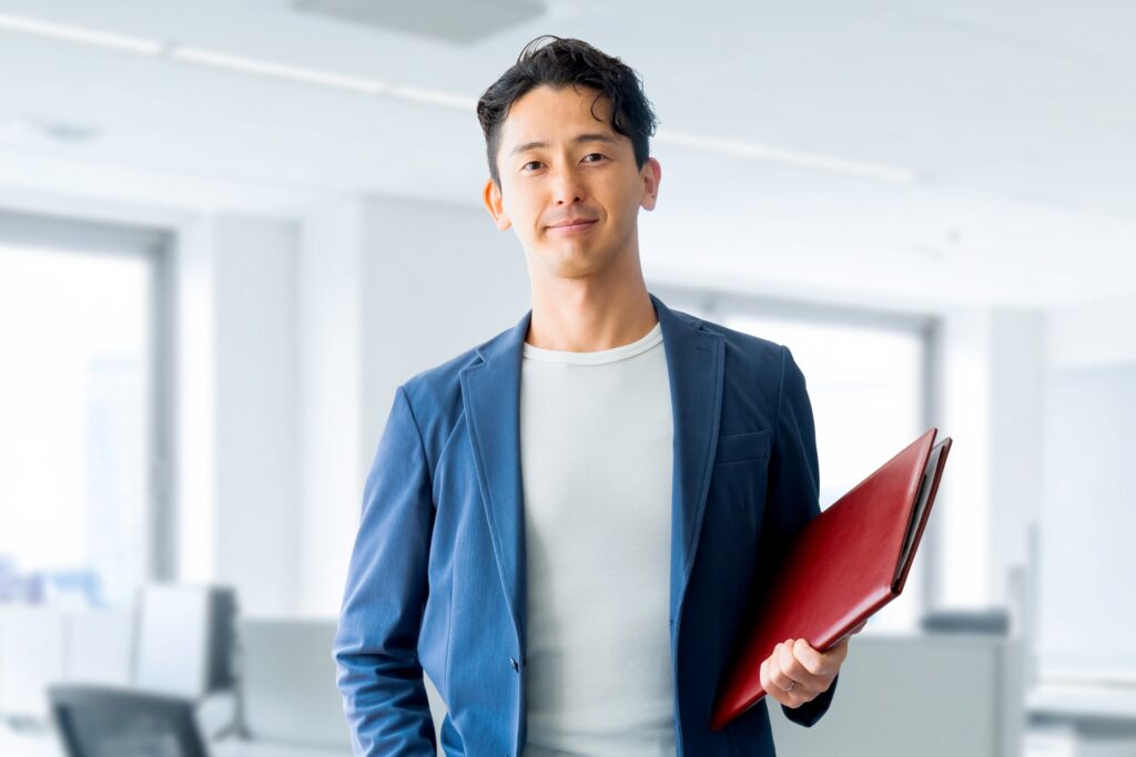 企業と対等に渡り合う産業ケアマネの新しい働き方。明るいオフィスで赤いファイルを持ち微笑む男性